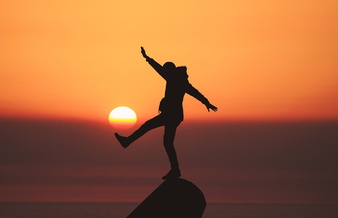 A person balances on one foot on a rock, arms outstretched, silhouetted against an orange sunset over the sea